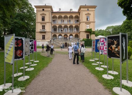 fotografia. art piknik w willi decjusza. ujęcie budynku willi od frontu. po dwóch stronach żwirowej alejki wystawa plakatów teatralnych, pomiędzy nimi spacerują goście.